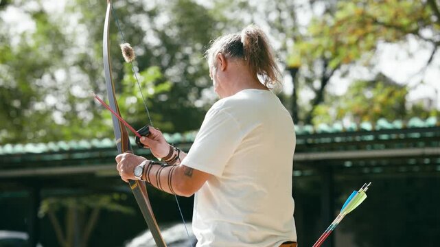 Focused mature archer with long hair pulling back the bowstring and releasing an arrow towards a target. Traditional archery practice outdoors