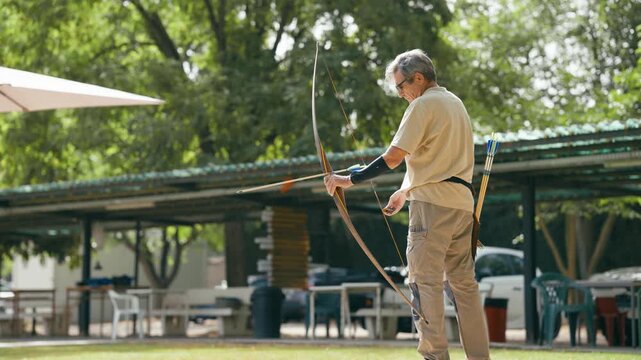 Mature man aiming and shooting a wooden bow. Active senior enjoying his hobby outdoors on a sunny day, focusing on precision and technique