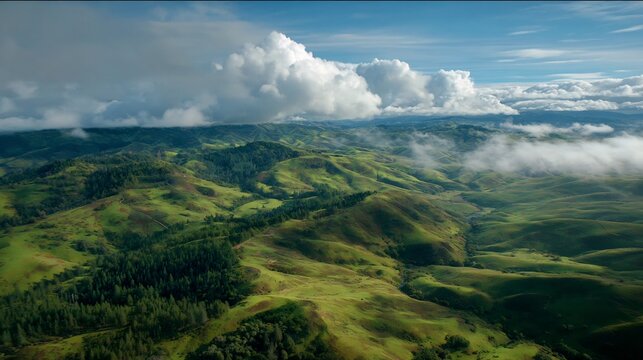 Aerial view of lush green rolling hills and valleys under a dramatic blue sky with fluffy white clouds and patches of mist, showcasing natural beauty and serene landscape.