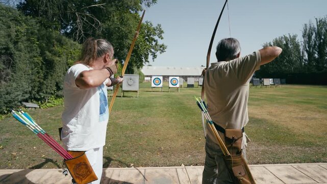 Two senior archers practicing at an outdoor archery range. One man shoots an arrow while the other observes his shot before shooting himself