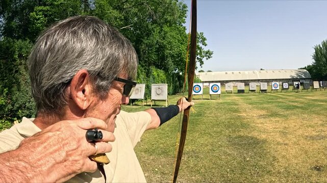 Senior man shooting an arrow with a traditional bow. Archer wearing sunglasses aiming at the target on a sunny day at an outdoor range