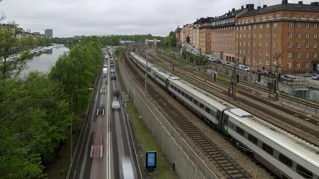 Cityscape timelapse of Stockholm&rsquo;s Klarastrandsleden with blurred cars, a long stationary train and many passing trains, and boats along the river, showing multiple transport modes.