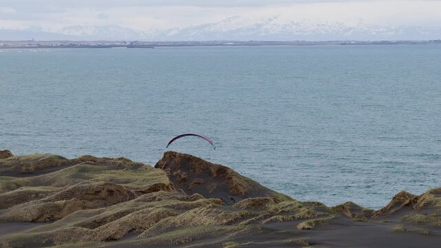 Paraglider flies low above weathered volcanic cliffs beside the Atlantic Ocean in southwest Iceland, with distant snow covered mountains across the water during a lateral drone track.