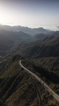 Dramatic aerial view following a curvy asphalt road along a steep mountain ridge in taiz, yemen, with a hazy valley and distant villages nestled among the rugged, sunlit peaks. In taiz yemen