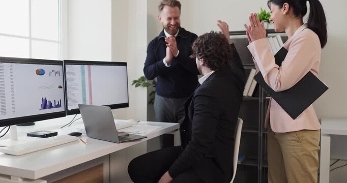 Teamwork colleagues celebrate success with applause in office. They cheer near monitors and a laptop as results drive business victory. Modern office. Teamwork achievement and motivation concept.