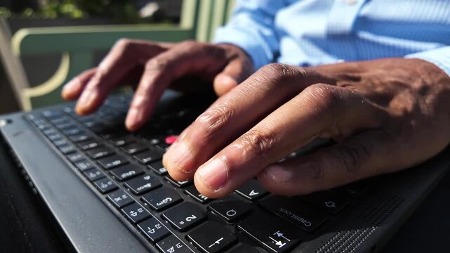 Hands of a black man rapidly typing on a laptop keyboard in slow motion under bright outdoor light.