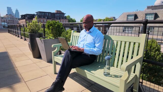 African american entrepreneur typing on a laptop and drinking water, sitting on a bench on a rooftop terrace with a city view in the background