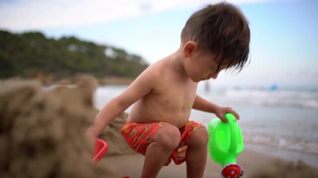 A toddler stands up and crouches again while playing with a watering can beside a sandcastle, with the sea in the background