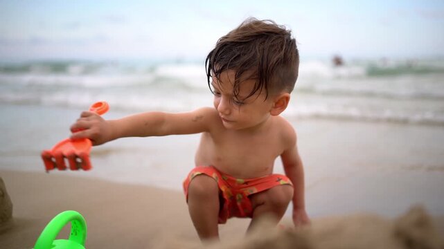 A boy crouches at the beach and plays with an orange rake as the sea stretches out behind him