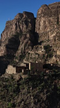 Impressive aerial view revealing an ancient, remote stone village perched precariously on a steep, rocky cliffside in the mountainous region of taiz, yemen, under a clear blue sky. In taiz yemen