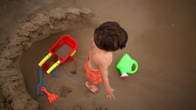 An overhead shot shows a boy tossing sand as waves wash in and out of a hole he dug at the shoreline