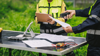 Fototapeta premium Engineers in safety vests review plans and use a radio beside solar panels and electrical testing tools at an outdoor renewable energy site.
