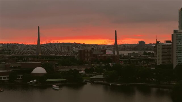 Subtle aerial rise of Boston's Zakim Bridge beneath a fiery orange dawn sky, with the Charles River, waterfront buildings, and a passing airplane in the far distance landing at Logan Airport