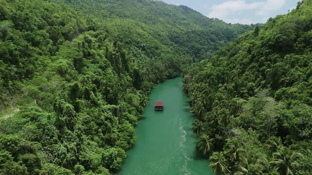 Drone pulls back and ascends as a Loboc River cruise boat travels along the green river between forested hills in Bohol