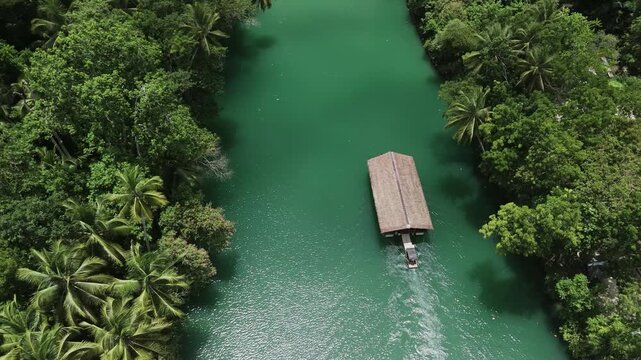 Aerial top-down view tilts up as a Loboc River cruise boat travels downstream through green water and forested hills in Bohol