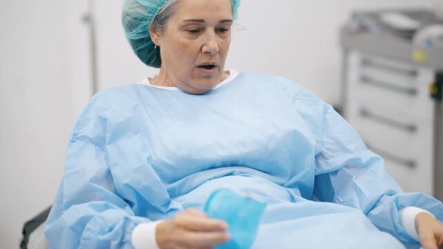 Anxious mature woman in a disposable gown and cap waking up from anesthesia after a surgical operation, feeling disoriented and distressed