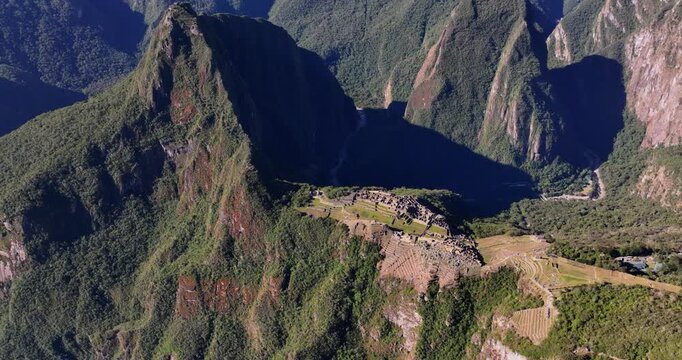 Aerial view of the Historic Sanctuary of Machu Picchu nestled among lush green Andean mountains and steep terraced slopes under bright sunlight in Urubamba Province, Cusco, Peru.