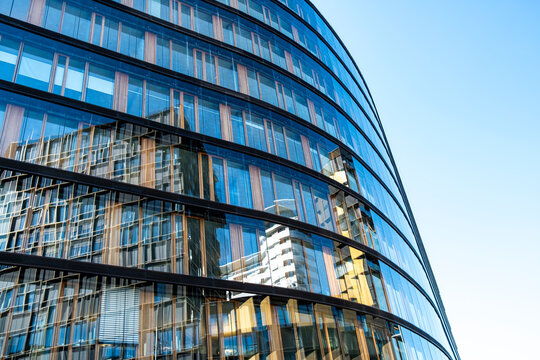 Curved blue glass facade with reflective windows and layered reflections creating a geometric pattern across modern urban architecture under clear daylight