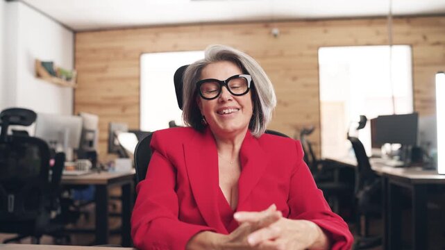 Cheerful senior businesswoman with gray hair and glasses having a video conference call, looking at the camera from the computer's point of view