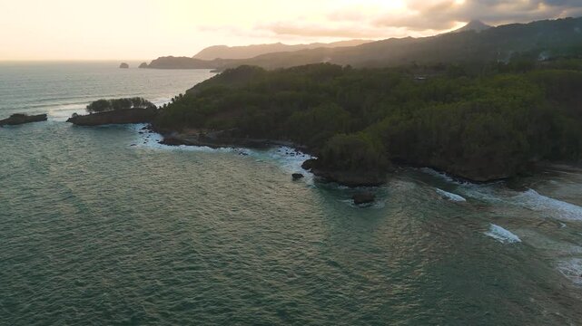 Aerial view of the rugged coastline with lush green cliffs and ocean waves crashing against the shore during sunset in Pacitan, East Java, Indonesia.