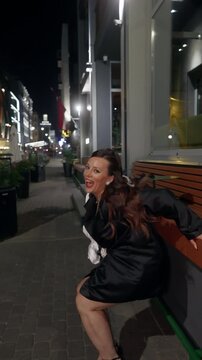 Elegant woman sitting on outdoor bench at night beside modern cafe walkway in city street