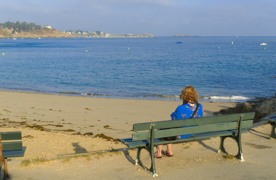A lone woman sits on a green bench facing the quiet Dinard beach, looking out toward the calm blue sea and the distant coast under the soft afternoon sky.