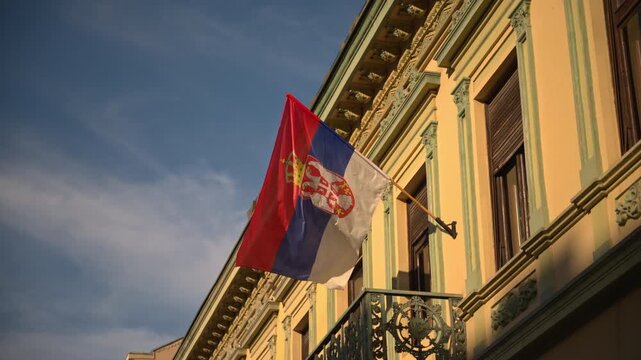 Serbian tricolor flag with coat of arms waves from a decorative balcony on a historic city building at sunset, evoking national pride, heritage architecture and urban atmosphere. 4K.