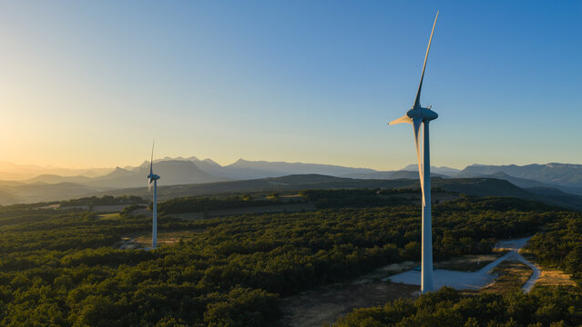 Aerial view of wind turbines rising above a forested landscape at golden sunrise, with layered mountain ranges stretching across the horizon in Marsanne, Auvergne-Rhone-Alpes, France.