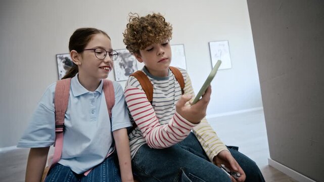 Caucasian children sitting in art gallery, looking at smartphone screen and pointing at device. Footage is suitable for educational apps, museum guide services, or youth technology marketing