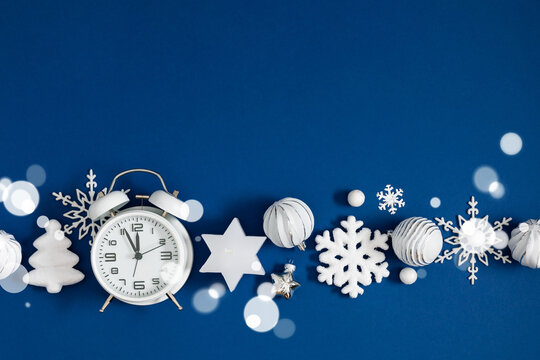 Christmas flat lay banner with white vintage alarm clock, snowflakes, and bauble ornaments on deep blue background with magical snow bokeh lights and large horizontal copy space for holiday text.