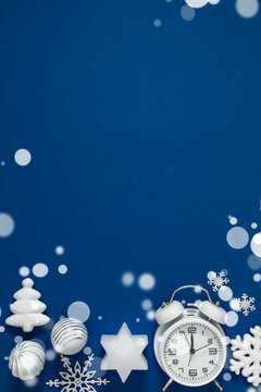 Vertical Christmas flat lay with white vintage alarm clock, snowflake ornaments, and star on deep blue background with magical snow bokeh lights and elegant copy space for holiday text.