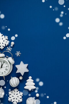 Vertical Christmas flat lay with white alarm clock, snowflakes, tree ornament, and baubles on a deep blue background with magical snow bokeh lights and large elegant copy space on the right side.