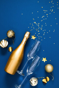 Luxury New Year celebration vertical flat lay with golden champagne bottle, flutes, baubles, and sparkling star confetti explosion on deep blue background with elegant creative copy space for text.