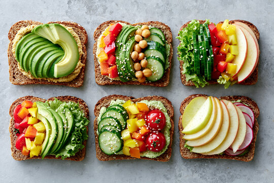 Six slices of bread topped with fresh, colorful ingredients: avocado, cucumbers, cherry tomatoes, hummus, chickpeas, bell peppers.