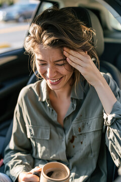 A driver sitting in a modern car, looking down with an amused, face-palm expression at a few drops of spilled coffee on their shirt.