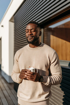 A man holding a white mug, standing on an outdoor deck in bright sunlight. He has his eyes closed and wears a beige crewneck sweater.