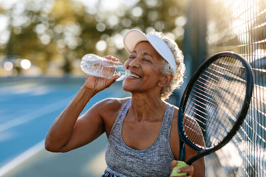 An elderly woman stands on a tennis court, smiling with closed eyes as she drinks water from a plastic bottle.