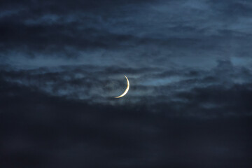 Thin waxing crescent moon shining through dark, layered clouds in a twilight sky. © thomasgweber