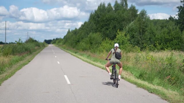 Highly engaging commercial ready video from behind of a funny young woman riding a bicycle on an empty countryside road, spreading her legs wide while pedaling.