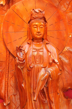 Close-up of a carved wooden Guanyin statue showing Vitarka mudra and holding a sacred vase against a carved lotus leaf backdrop.