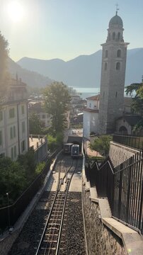 Funicular Ascending in European Town: A steep funicular railway track winds through a charming European town, with historic buildings and a bell tower overlooking a serene lake. Switzerland, Lugano