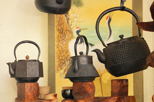 Vintage cast iron teapots and a hanging tetsubin kettle against a traditional oriental painting with cranes.
