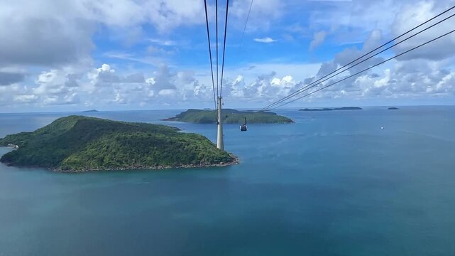 Aerial view of a cable car traveling over green hills and turquoise waters, with distant islands visible under blue sky. The world's longest cable car system, connecting Phu Quoc with Hon Thom island