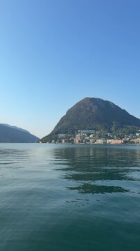 Mountain and Lake Vista: A majestic mountain range rises above a tranquil lake under a clear blue sky, Switzerland, Lugano, Lake Lugano