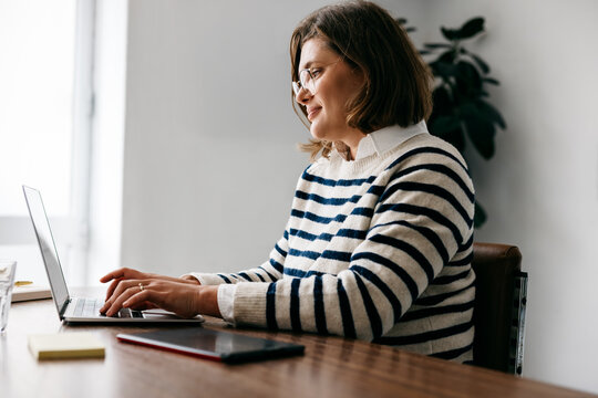 Woman working on laptop at desk in office with smartphone and notebook