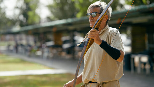 Older man holding a traditional recurve bow, focusing on his shot and aiming at a target during an archery practice session