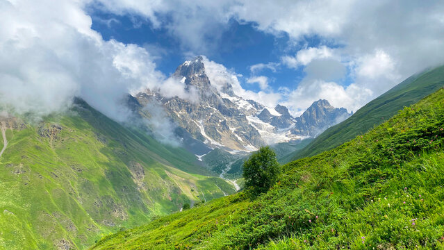Clouds part around Ushba, wide view from a green alpine valley in Svaneti, Georgia, presenting the mountain approach landscape