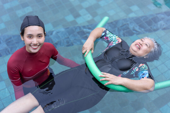 A smiling caregiver supports a senior woman floating with a pool noodle during an aquatic therapy session in an outdoor swimming pool. The image represents rehabilitation, trust, and healthy aging