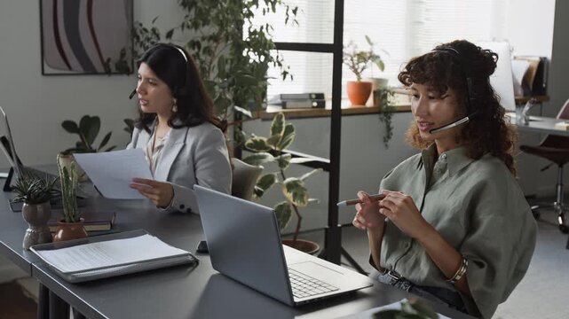Caucasian and Hispanic young adult female colleagues wearing headsets working in office, talking to clients and analyzing documents. Ideal for customer support or corporate videos