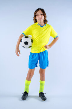 Male youth soccer player with long hair wearing yellow and blue uniform standing with soccer ball under one arm